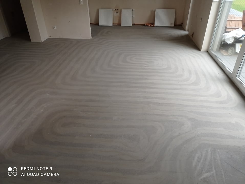 Świeżo wylana, szara posadzka z widocznym układem rur systemu grzewczego w kształcie meandrów, przygotowana do dalszych prac wykończeniowych; w tle widoczne grzejniki naścienne.