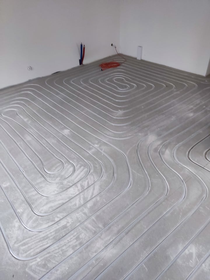 Rury instalacji grzewczej ułożone w pętle na podłodze w surowym stanie, widoczne wystające rury instalacyjne na ścianie.