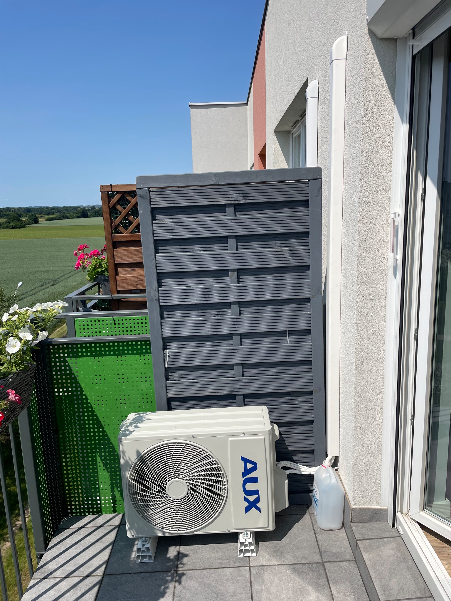 Biała jednostka zewnętrzna klimatyzacji marki AUX na balkonie z szarą osłoną i widokiem na zielone pola w tle.