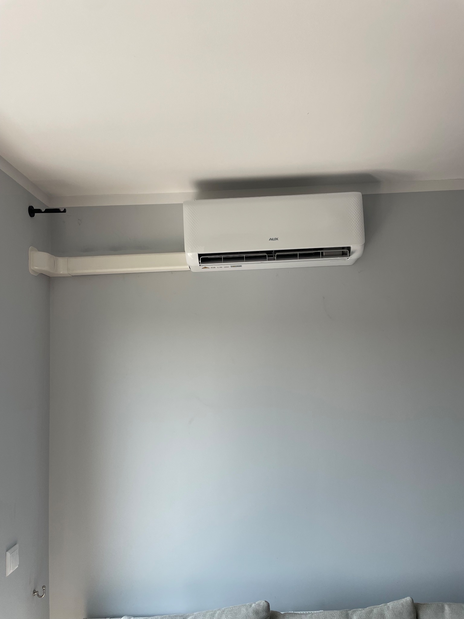 Biała klimatyzacja AUX zamontowana na szarej ścianie w pomieszczeniu, z widoczną instalacją prowadzącą wzdłuż ściany.