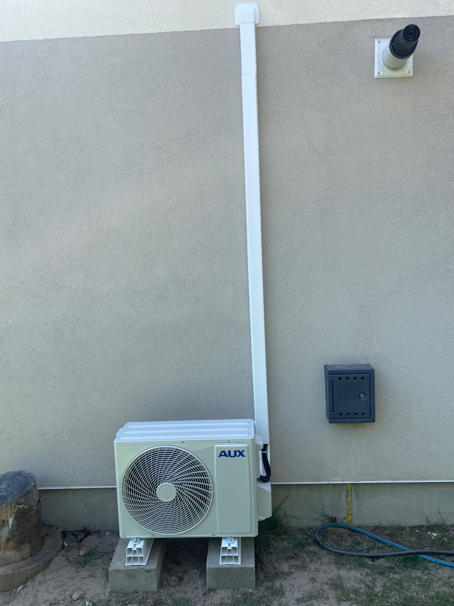 Klimatyzator AUX zamontowany na zewnątrz budynku, z widoczną instalacją odprowadzającą. Obok skrzynka gazowa i komin. Ściana w jasnym odcieniu beżu.