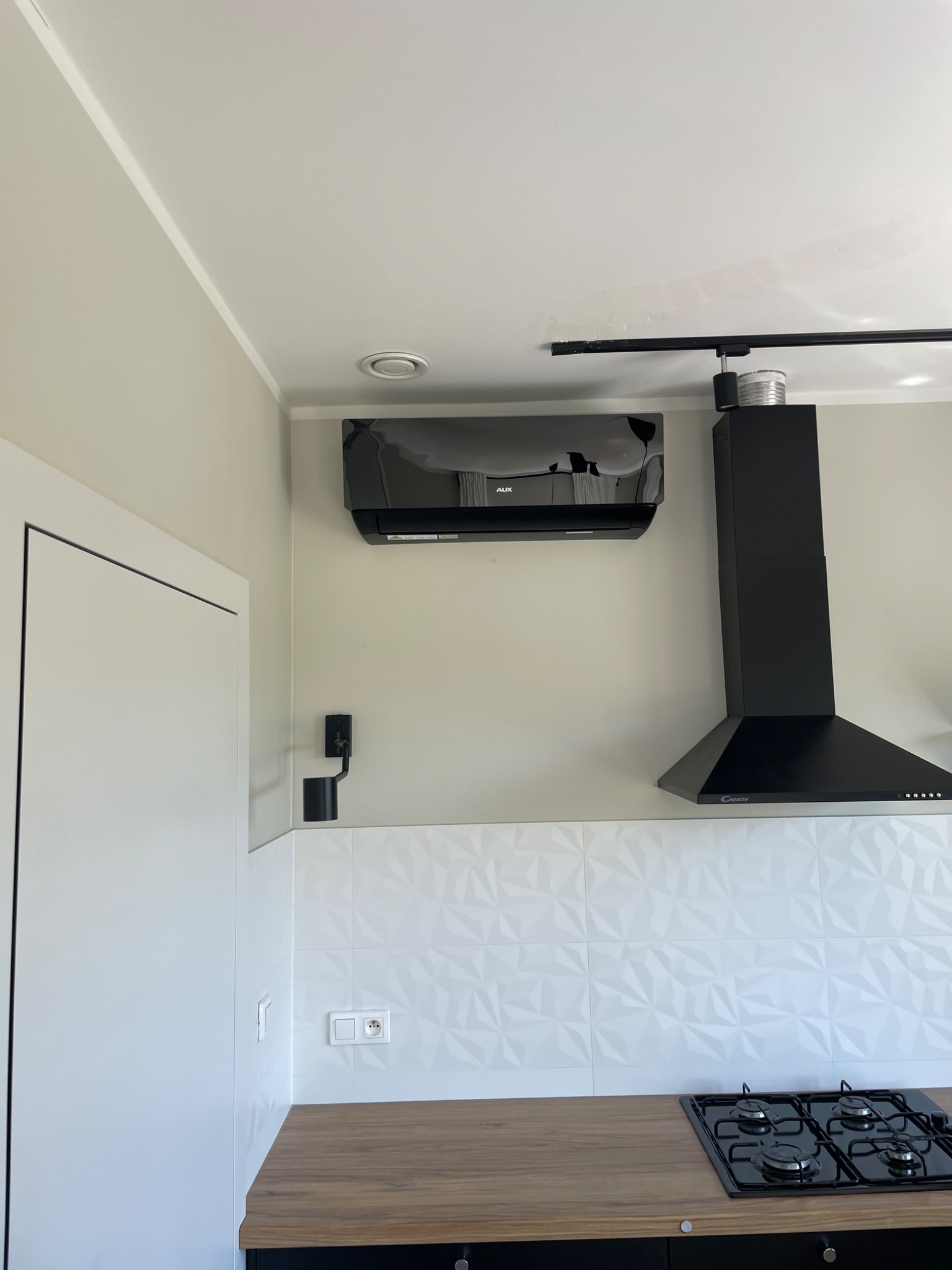 Czarna klimatyzacja AUX nad płytą grzewczą i okapem Candy w nowoczesnej kuchni z białymi ścianami i drewnianym blatem. Widoczne oświetlenie sufitowe.