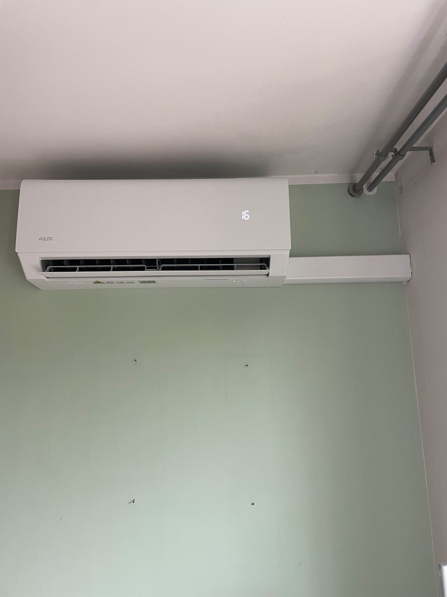 Biały klimatyzator ścienny AUX zamontowany na zielonej ścianie, z widoczną temperaturą 16 stopni Celsjusza. Rury instalacyjne w rogu pomieszczenia.