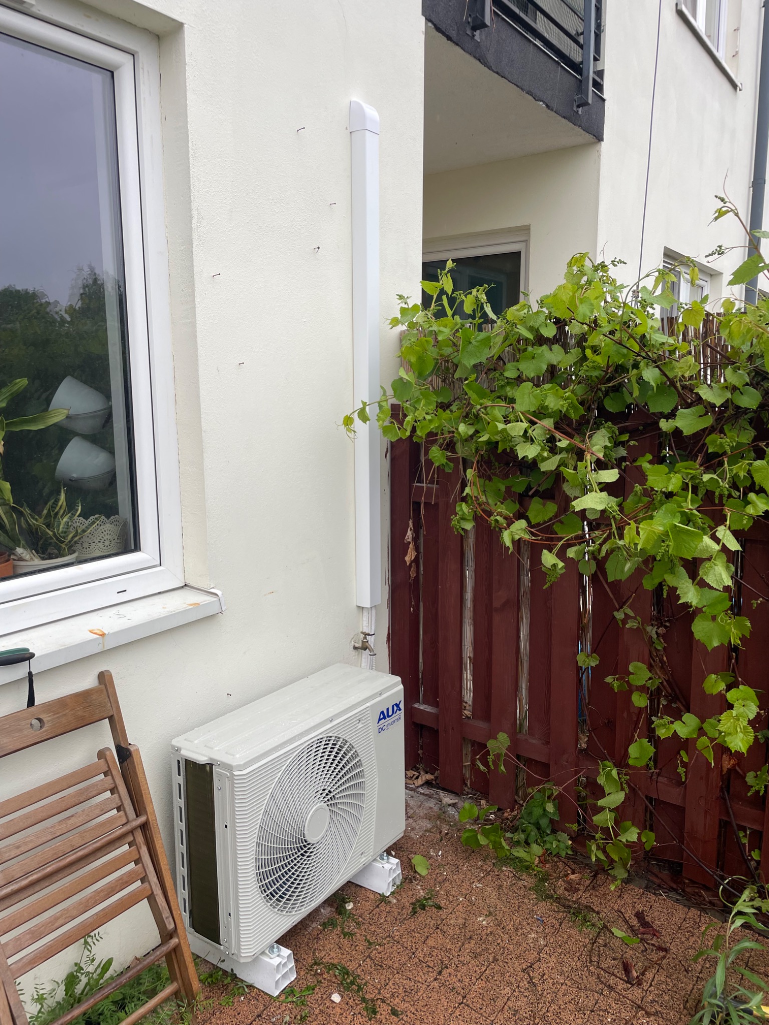 Biały klimatyzator AUX zamontowany na zewnątrz budynku, obok drewnianego płotu z pnącymi roślinami i składanym krzesłem. Widoczna biała rura odprowadzająca.
