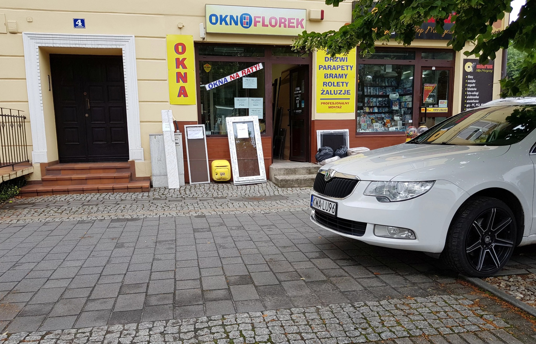 Sklep z oknami i drzwiami 'Okno Florek': wystawione przy wejściu okna i drzwi, obok zaparkowany biały samochód Skoda Superb z widoczną tablicą rejestracyjną.