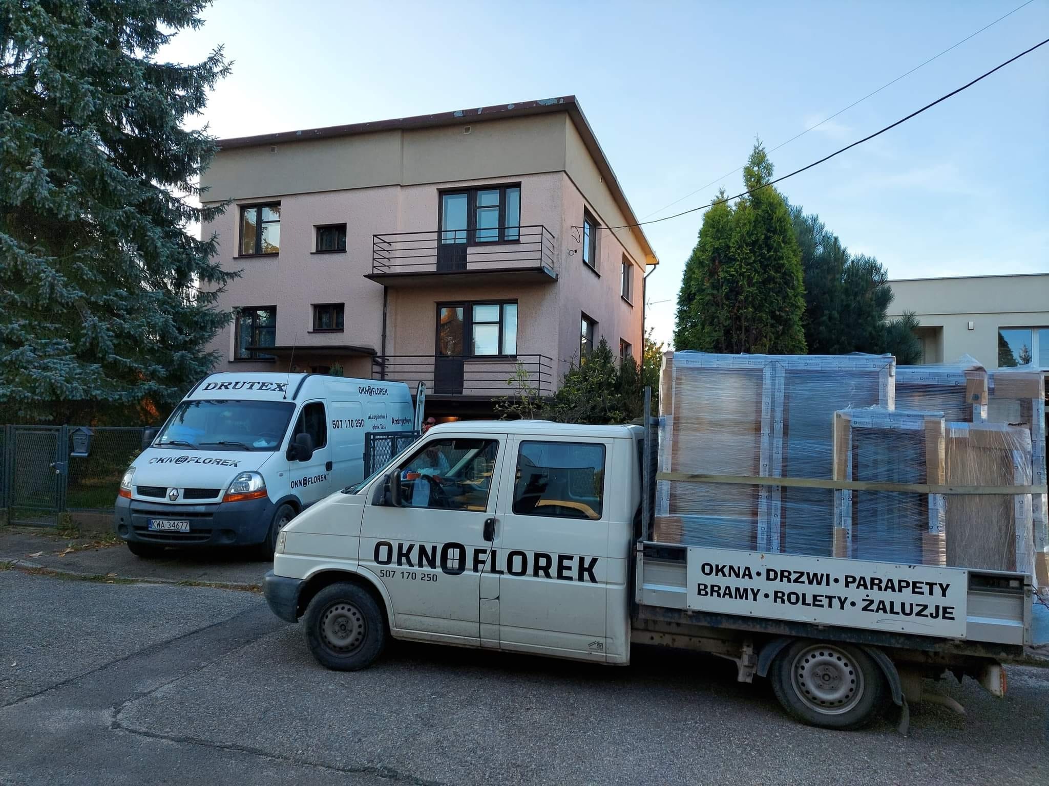 Dwa busy firmy OknoFlorek, jeden załadowany oknami i drzwiami w folii, stojące na tle dwupiętrowego budynku z balkonem.