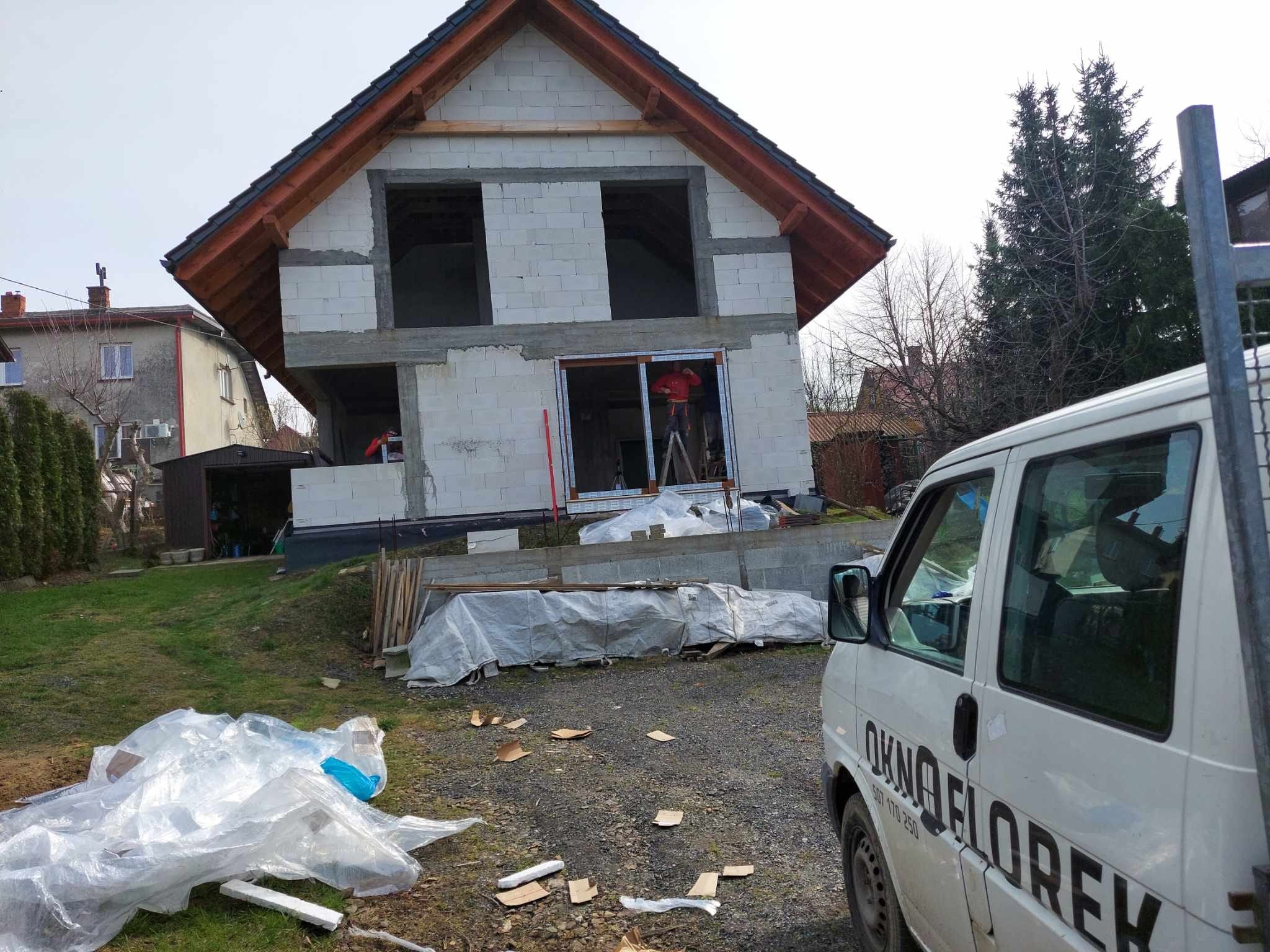 Budowa domu jednorodzinnego w stanie surowym otwartym, widoczny montaż okna przez pracownika na drabinie, na pierwszym planie biały bus z napisem 'OKNO FLOREK'.