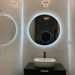 Elegancka łazienka z białymi marmurkowymi płytkami, okrągłym lustrem z podświetleniem LED i białą umywalką nablatową na czarnej szafce z frezowanymi frontami.