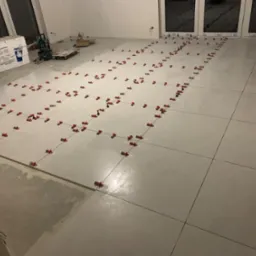 Układanie jasnych, kwadratowych płytek podłogowych z użyciem czerwonych klinów poziomujących; widoczny fragment surowej posadzki i okna z białymi ramami.