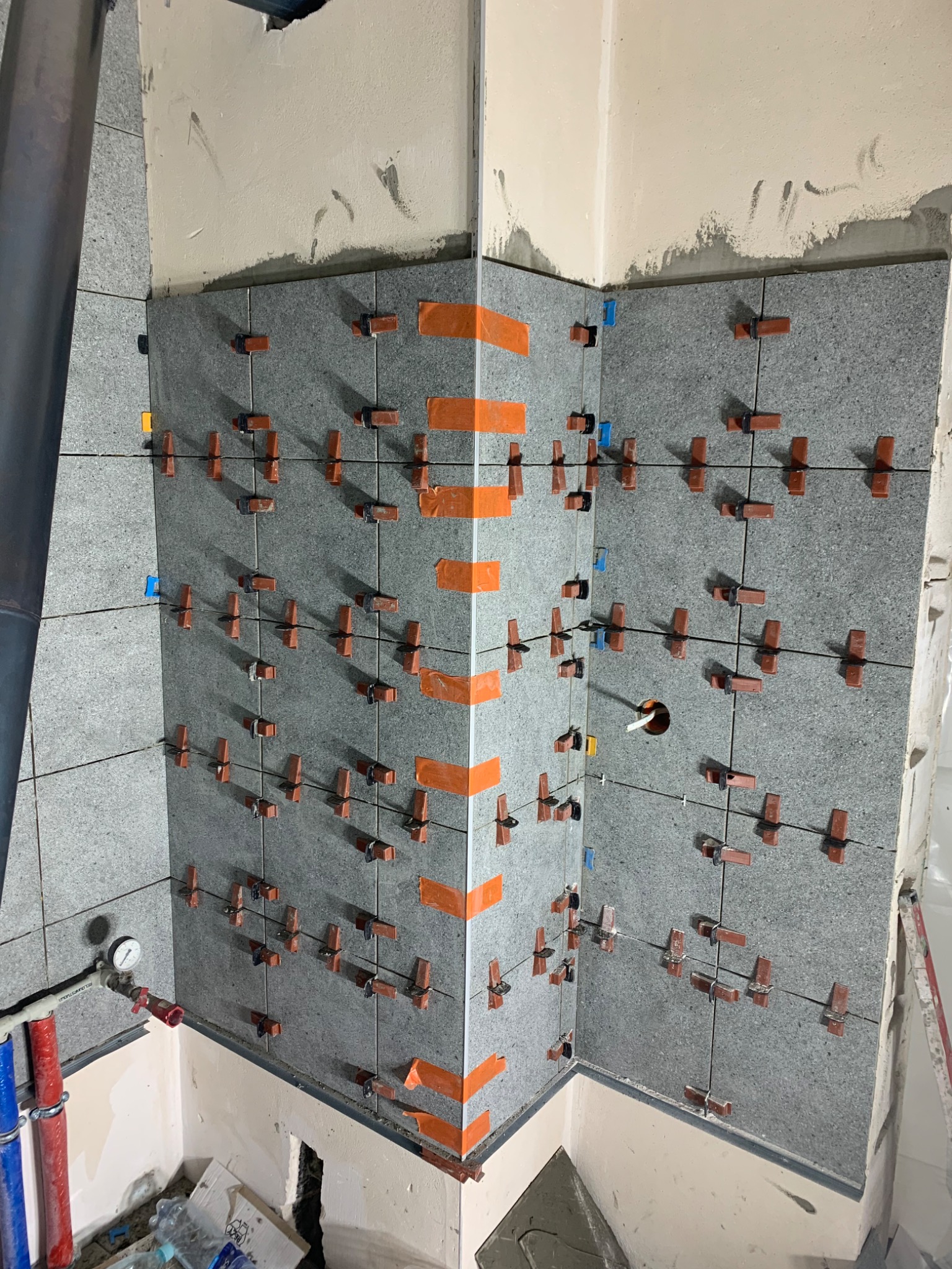 Świeżo ułożone szare płytki na ścianie w narożniku, zabezpieczone pomarańczową taśmą i brązowymi krzyżykami dystansowymi, widoczny fragment instalacji wodnej z rurami w kolorze niebieskim...