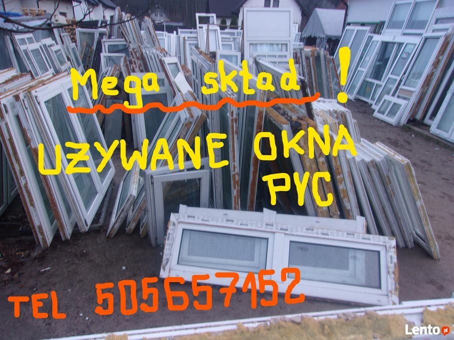 Składowisko używanych okien i drzwi, widoczne białe ramy okienne różnej wielkości, część z nich oparta o siebie pod kątem, na ziemi, w tle budynki mieszkalne.