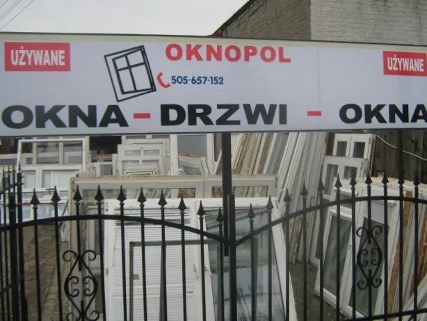 Szyld firmy OKNOPOL z napisem OKNA - DRZWI - OKNA, widoczne używane okna za metalowym ogrodzeniem.