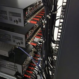 LVA Systems Aleksander Kociemba - Profesjonalna instalacja sprzętu audio-wideo w szafie rack, z rzędami czarnych urządzeń i gęstym, uporządkowanym okablowaniem z czerwonymi złączami i etykietami.