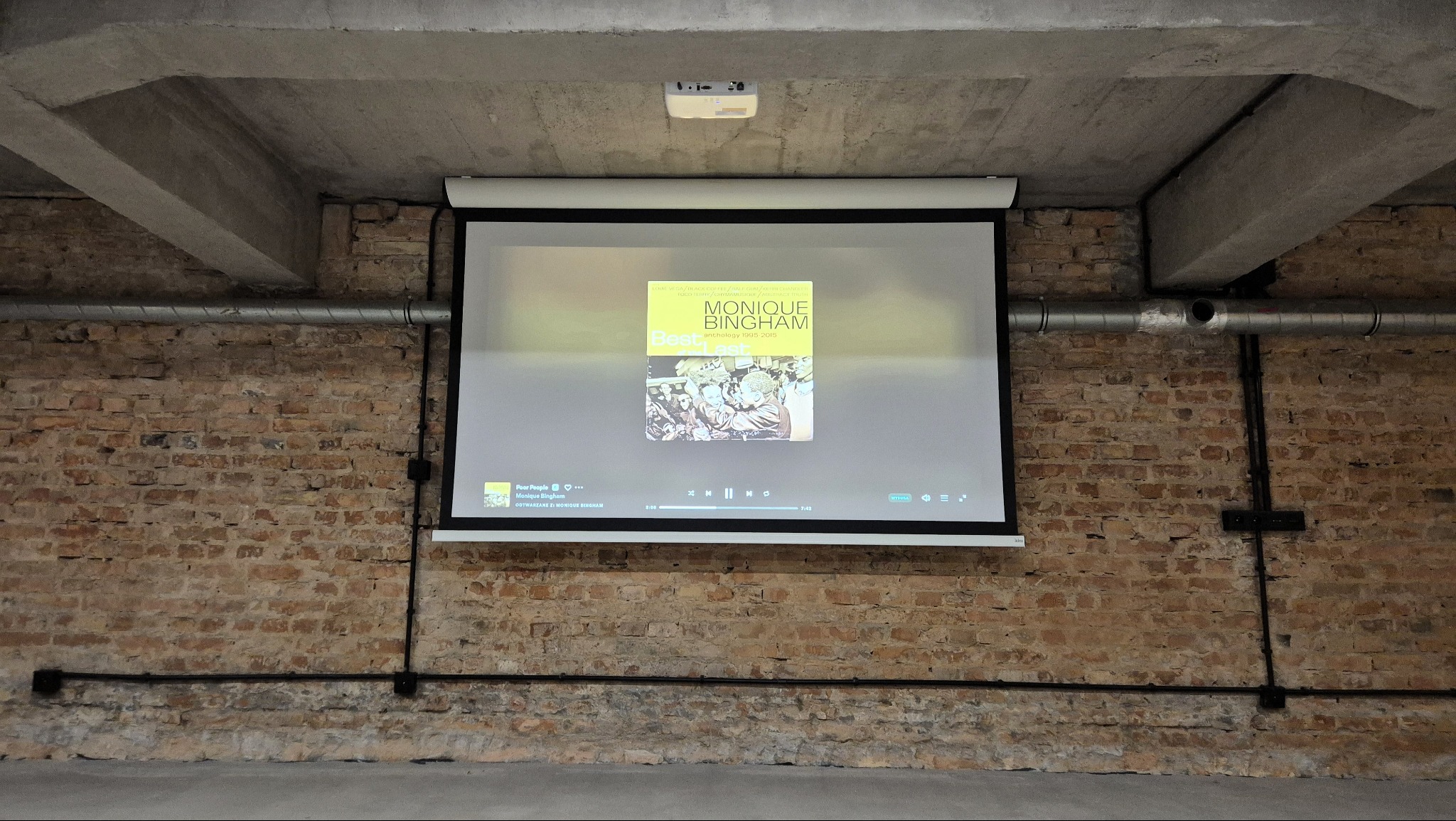Ekran projekcyjny z okładką albumu 'Monique Bingham' na tle ceglanej ściany. Sufitowy projektor, widoczne odkryte instalacje elektryczne i wentylacyjne na surowym betonie.