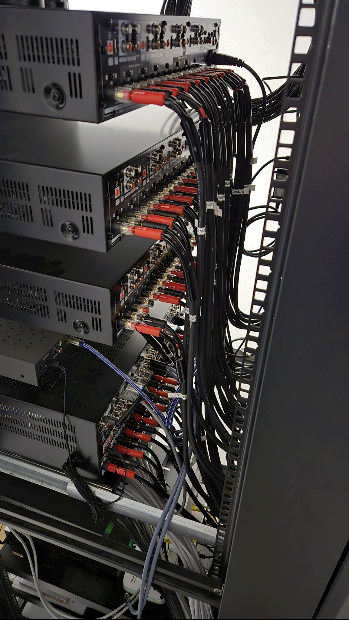 Profesjonalna instalacja sprzętu audio-wideo w szafie rack, z rzędami czarnych urządzeń i gęstym, uporządkowanym okablowaniem z czerwonymi złączami i etykietami.