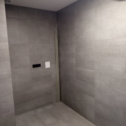 Szare, matowe płytki w surowej łazience, widoczne pionowe rury przy ścianie, czarna armatura i biała zaślepka. Minimalistyczny design, stonowana kolorystyka.