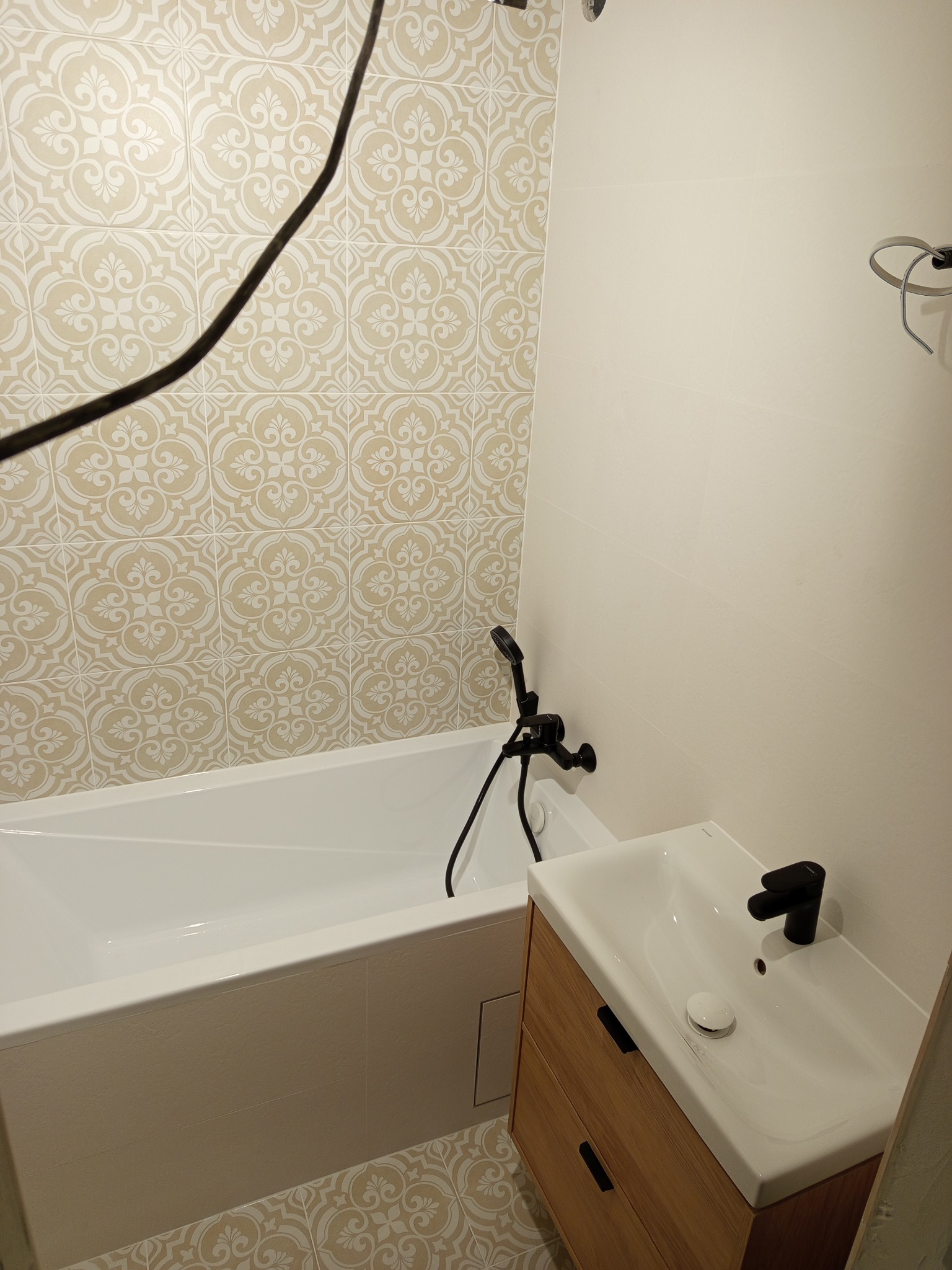 Wykończona łazienka z beżowo-białymi płytkami wzorzystymi na podłodze i ścianie, białą wanną i umywalką z drewnianą szafką oraz czarną armaturą.