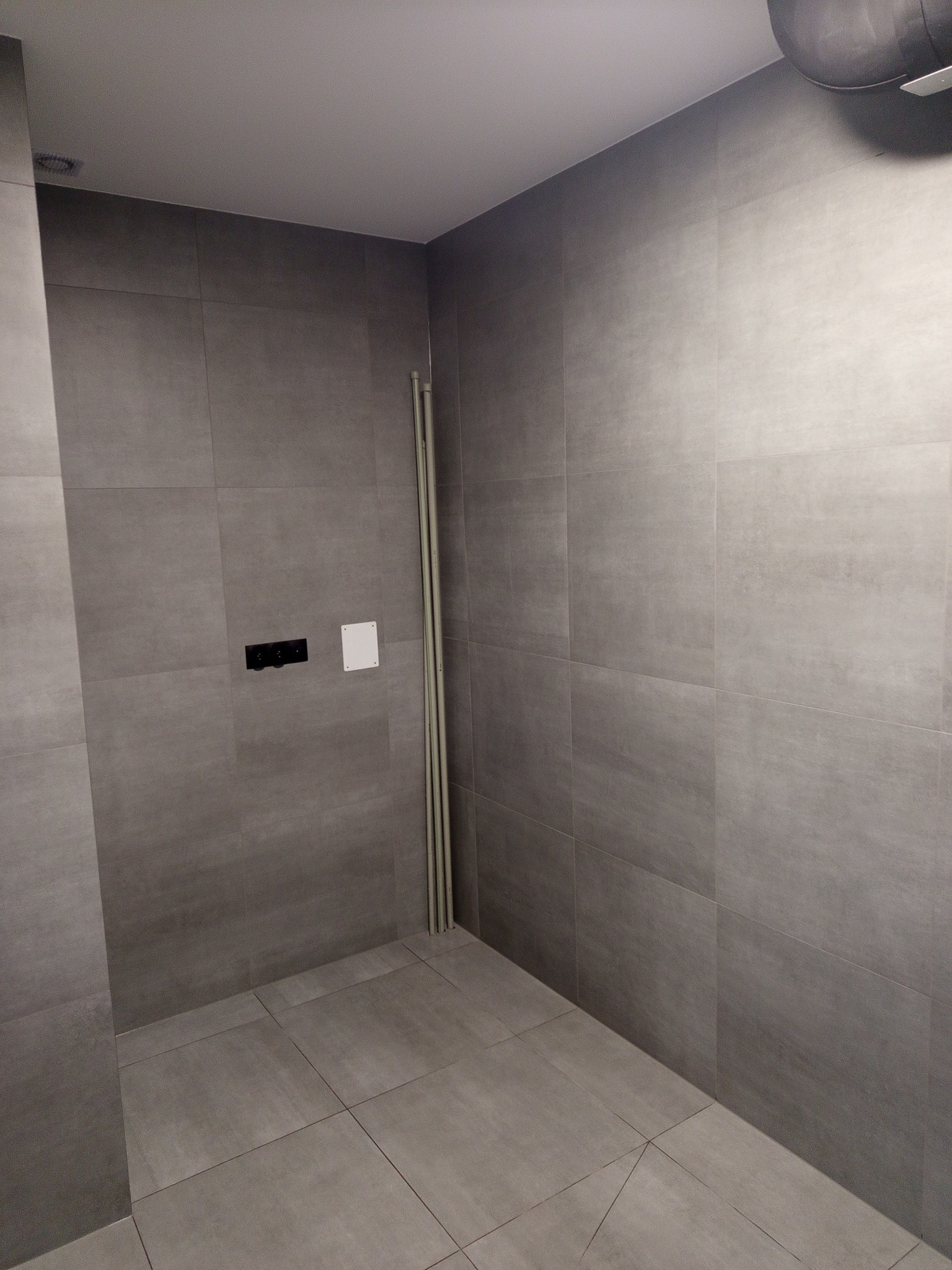 Szare, matowe płytki w surowej łazience, widoczne pionowe rury przy ścianie, czarna armatura i biała zaślepka. Minimalistyczny design, stonowana kolorystyka.
