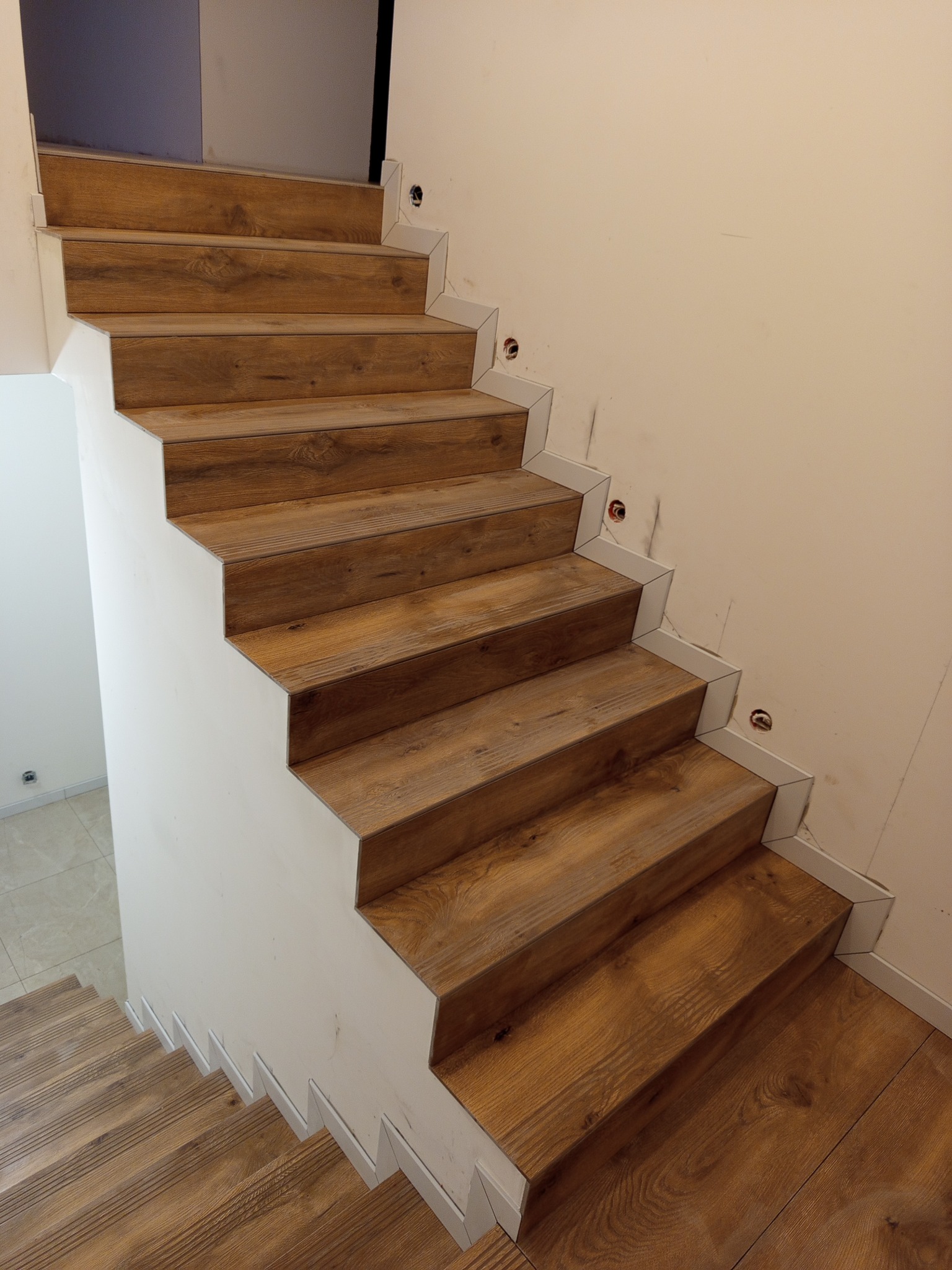 Schody wykończone panelami drewnopodobnymi z białymi listwami, widok z dołu. Widoczne niedokończone gniazdka elektryczne na ścianie.