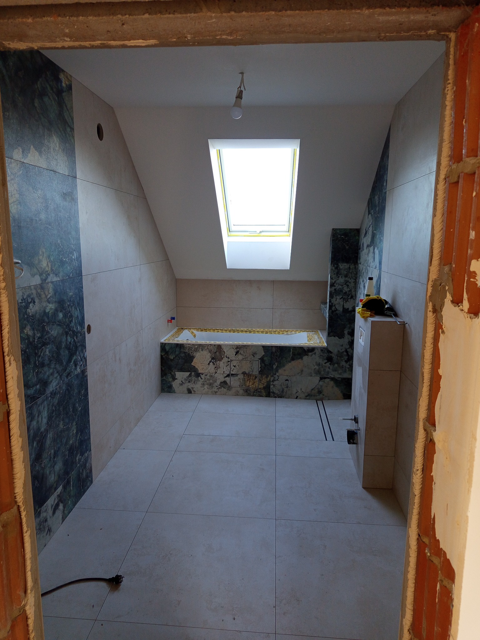 Wykończona łazienka na poddaszu z oknem dachowym. Jasne płytki podłogowe i ścienne, fragment ściany z ciemnym marmurem. Widok zza surowej framugi drzwi.