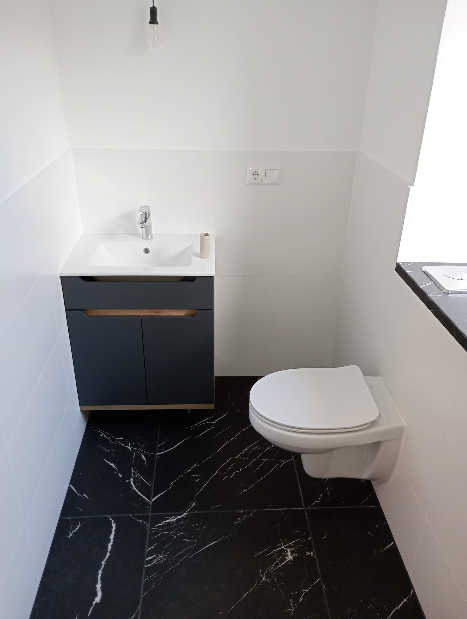 Nowoczesna łazienka z czarną marmurową podłogą, białymi płytkami ściennymi i wiszącą toaletą. Szafka pod umywalką w kolorze grafitowym z drewnianymi akcentami.