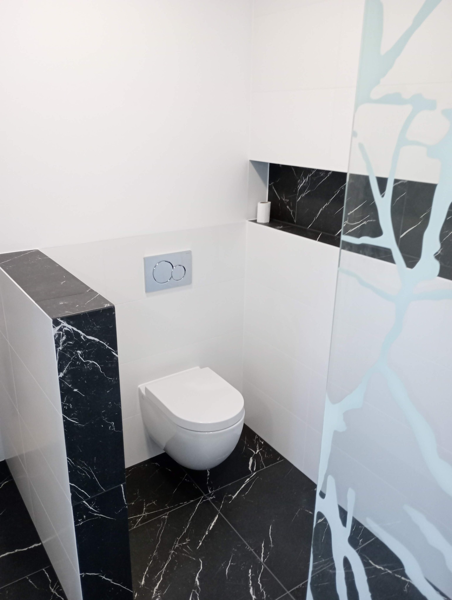 Nowoczesna toaleta z czarno-białymi płytkami imitującymi marmur, podwieszana miska WC i szklana ścianka z motywem drzewa.
