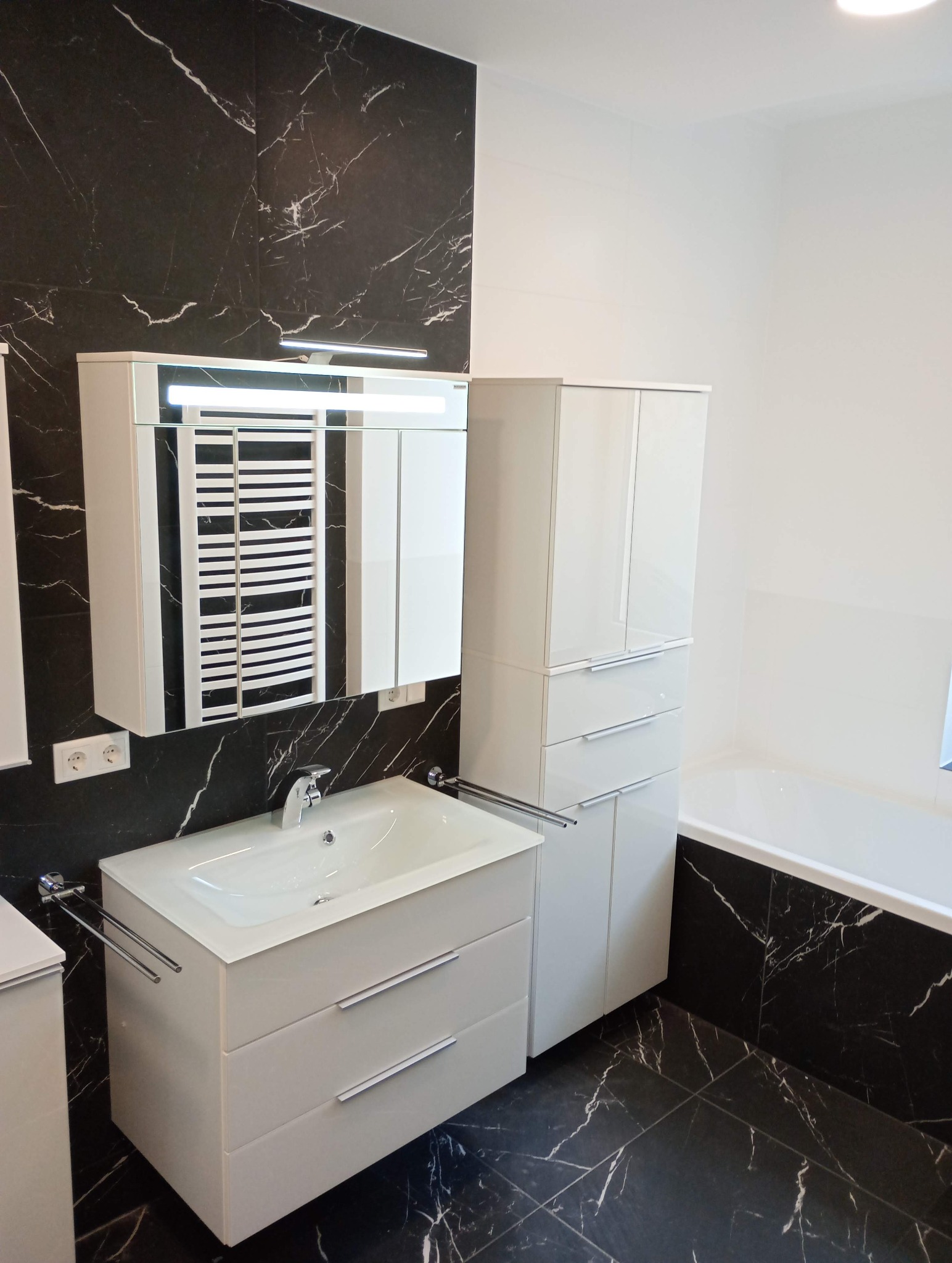 Wykończona łazienka z czarno-białymi płytkami imitującymi marmur, białą szafką z umywalką i lustrem z oświetleniem. Widoczna wanna i grzejnik drabinkowy.