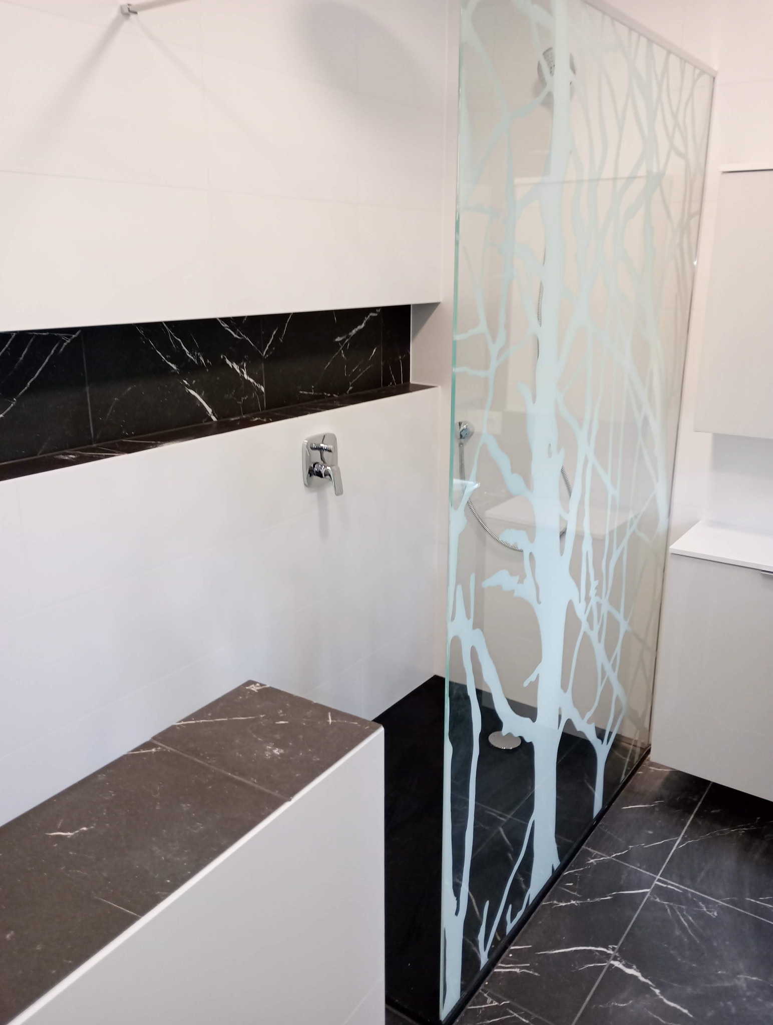 Nowoczesna łazienka z kabiną prysznicową ze szkła z motywem drzewa, czarno-białe płytki marmurowe na ścianach i podłodze, chromowana armatura.