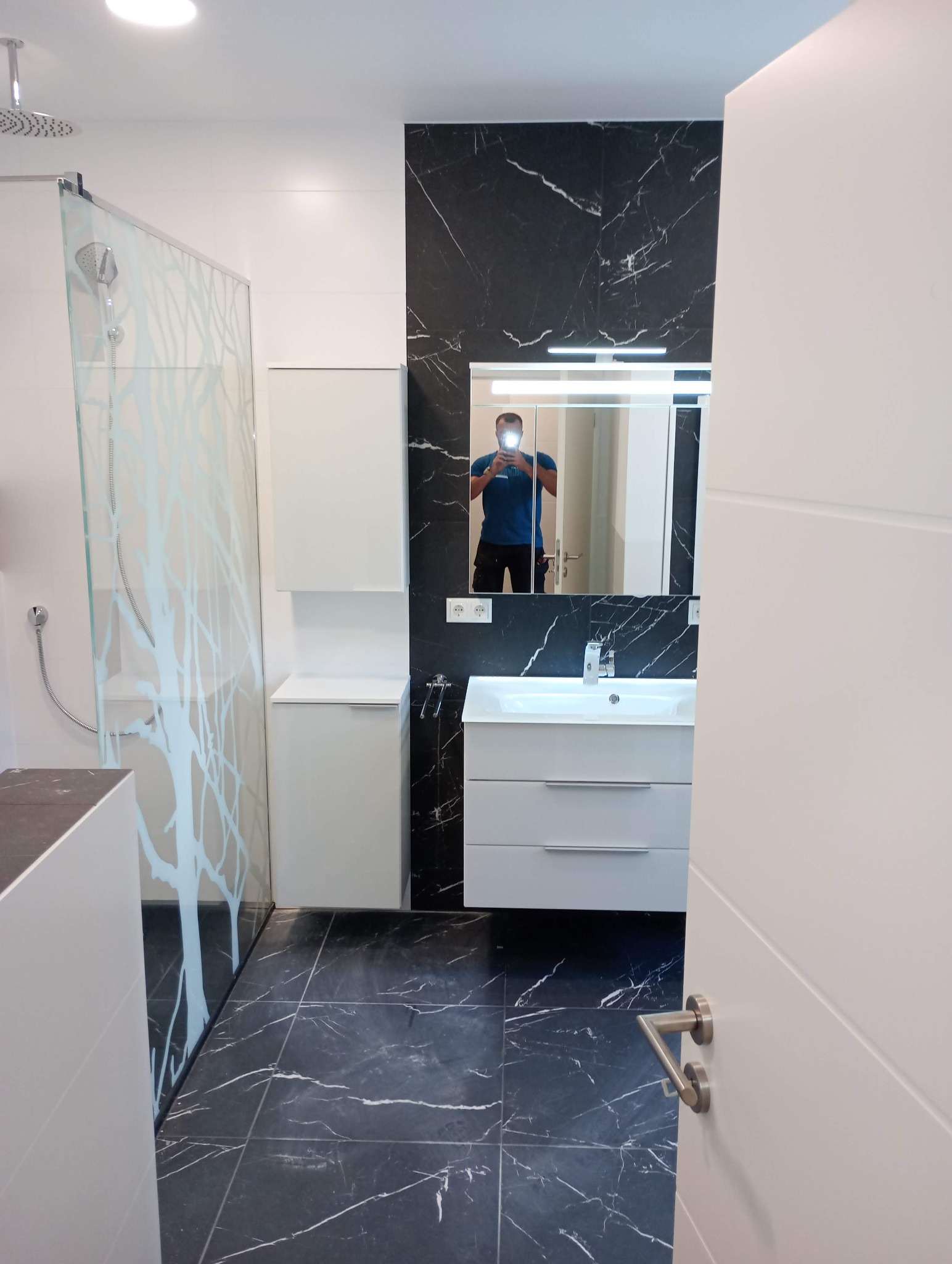 Nowoczesna łazienka z czarnymi płytkami imitującymi marmur, białą armaturą i kabiną prysznicową z motywem drzewa. Widoczne odbicie osoby robiącej zdjęcie w lustrze.