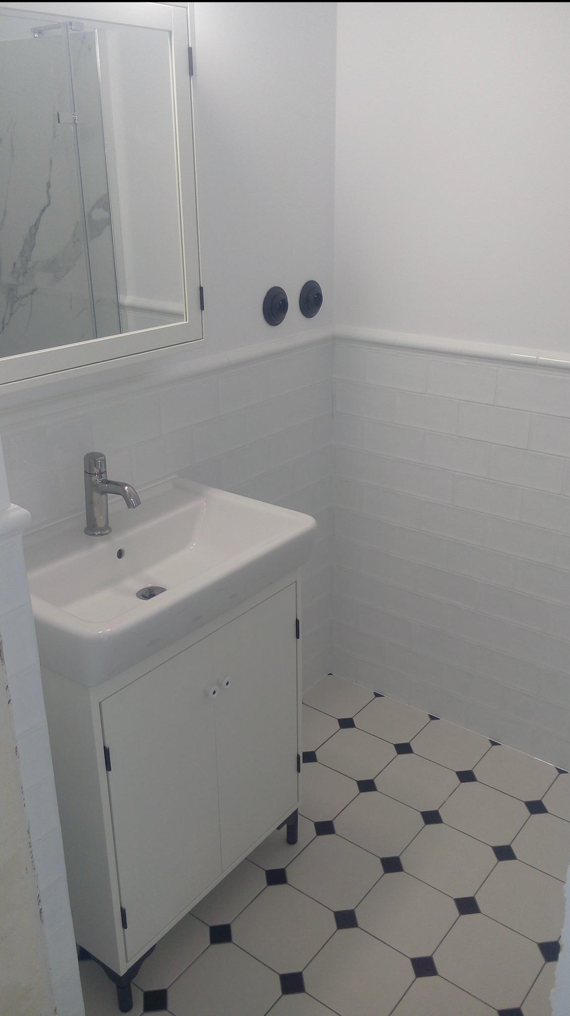 Jasna łazienka z białą umywalką na szafce, lustrem i czarno-białą podłogą w karo. Ściany wyłożone białymi, prostokątnymi płytkami. Widoczne czarne elementy armatury.