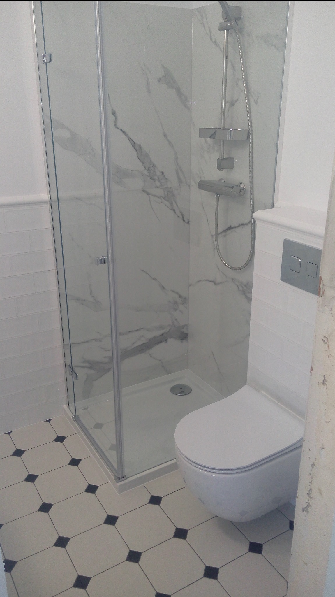 Nowoczesna łazienka po remoncie: kabina prysznicowa z marmurowym wzorem, biała toaleta i podłoga w biało-czarną szachownicę. Widoczne detale wykończenia.
