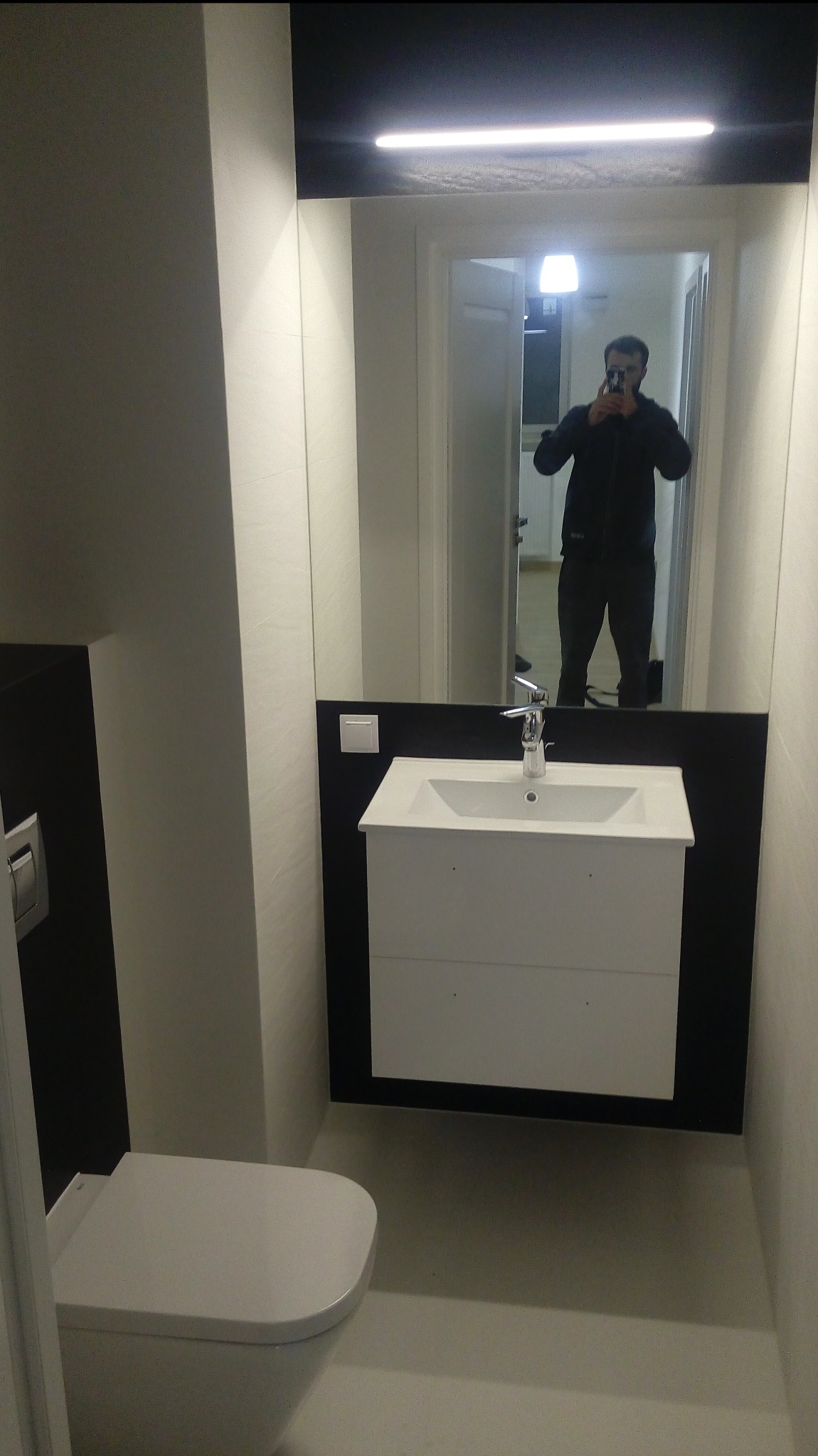 Nowoczesna, minimalistyczna łazienka w bieli i czerni. Umywalka z szafką, toaleta podwieszana, duże lustro z odbiciem osoby robiącej zdjęcie. Oświetlenie LED.
