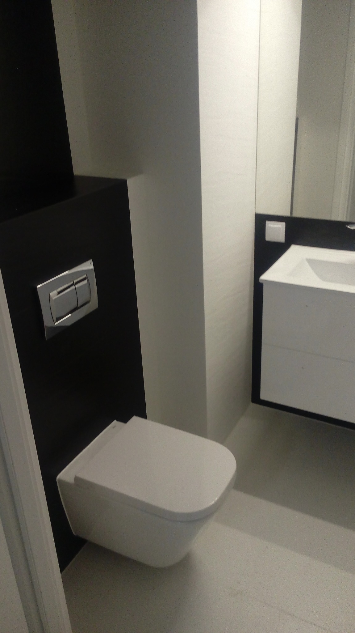 Nowoczesna, minimalistyczna łazienka z podwieszaną toaletą na czarnej ścianie i białą umywalką. Widoczne detale wykończenia i armatura.