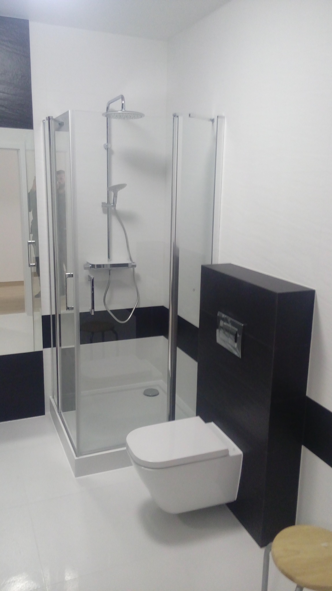 Nowoczesna łazienka z kabiną prysznicową i podwieszaną toaletą. Białe płytki kontrastują z czarnym pasem na ścianie. Widoczny fragment stołka.