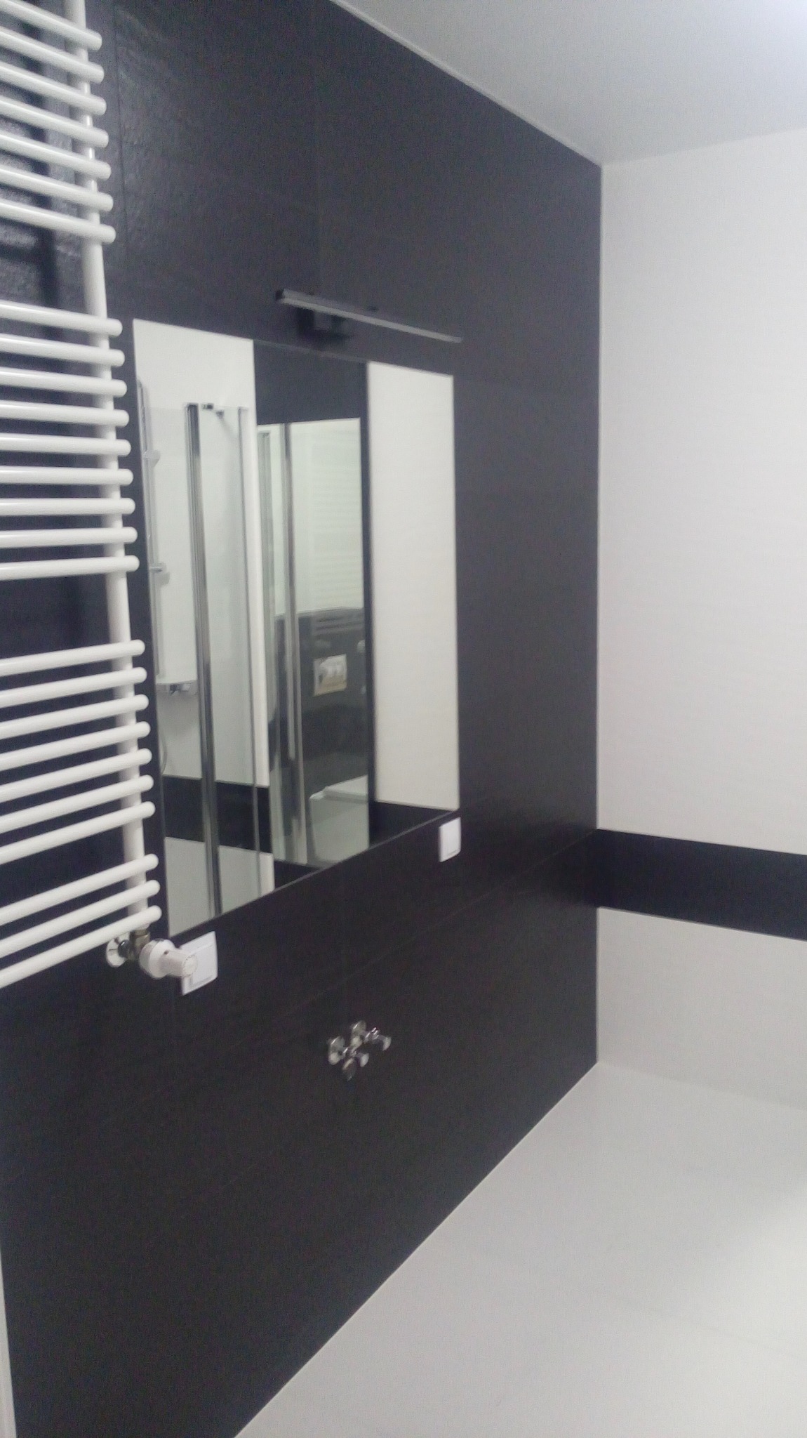 Nowoczesna łazienka w czarno-białej kolorystyce. Widoczny grzejnik drabinkowy, lustro z oświetleniem i kabina prysznicowa w odbiciu. Minimalistyczny design.