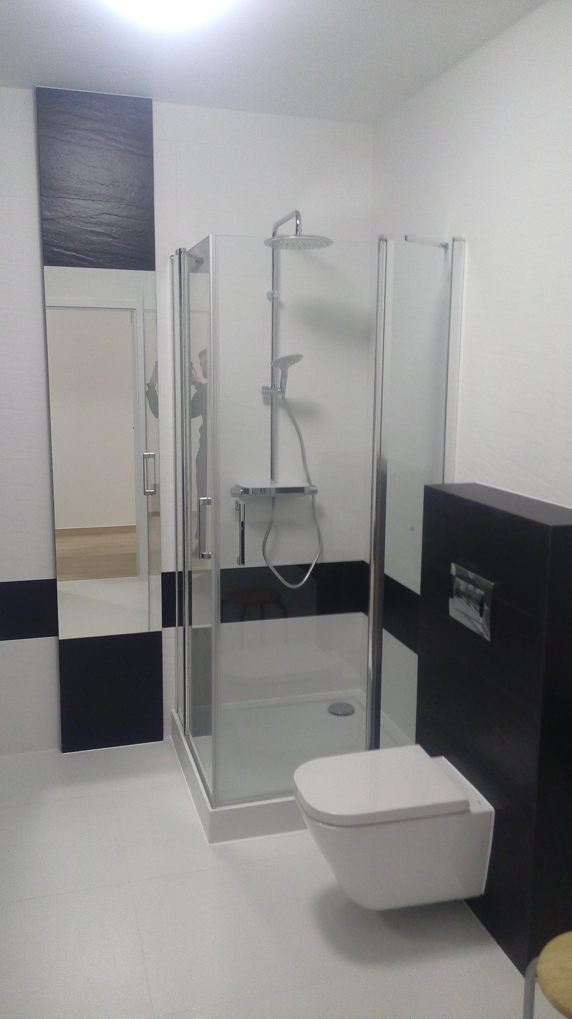 Nowoczesna łazienka z kabiną prysznicową typu walk-in, podwieszaną toaletą i czarno-białymi akcentami na ścianach. Minimalistyczny design, jasne płytki podłogowe.