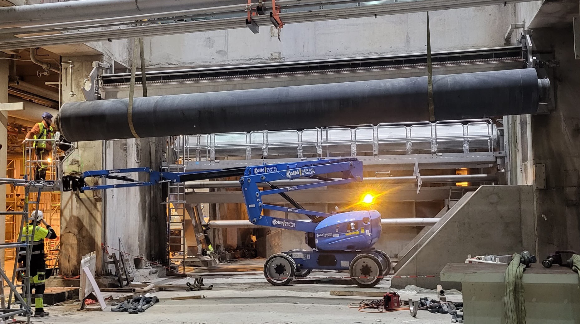 Montaż dużej, czarnej rury wewnątrz betonowej konstrukcji za pomocą podnośnika koszowego; pracownicy w odblaskowych ubraniach nadzorują proces.