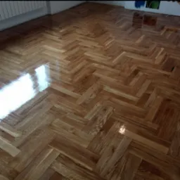 Parkiet ułożony w jodełkę, po renowacji z połyskiem, widoczne odbicie światła na powierzchni. W tle ściana z grzejnikiem i fragmentem malowidła.