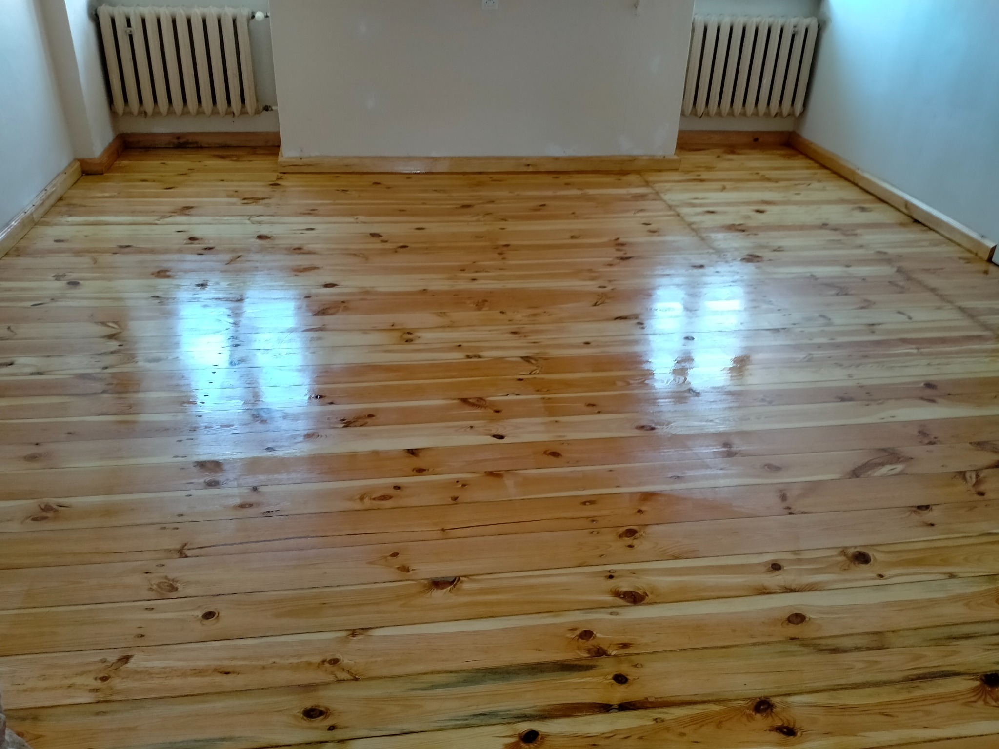 Wypolerowana, lakierowana podłoga z jasnego drewna w pokoju z białymi ścianami i grzejnikami.