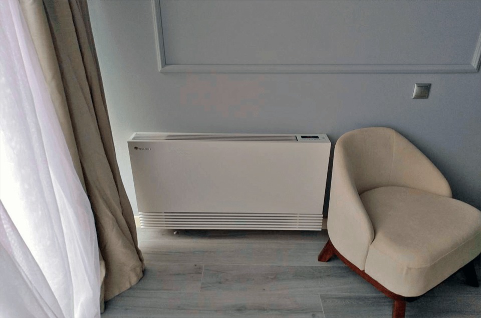 Biały rekuperator pokojowy z wyświetlaczem, umieszczony przy ścianie w jasnym pomieszczeniu, obok kremowego fotela i zasłony.