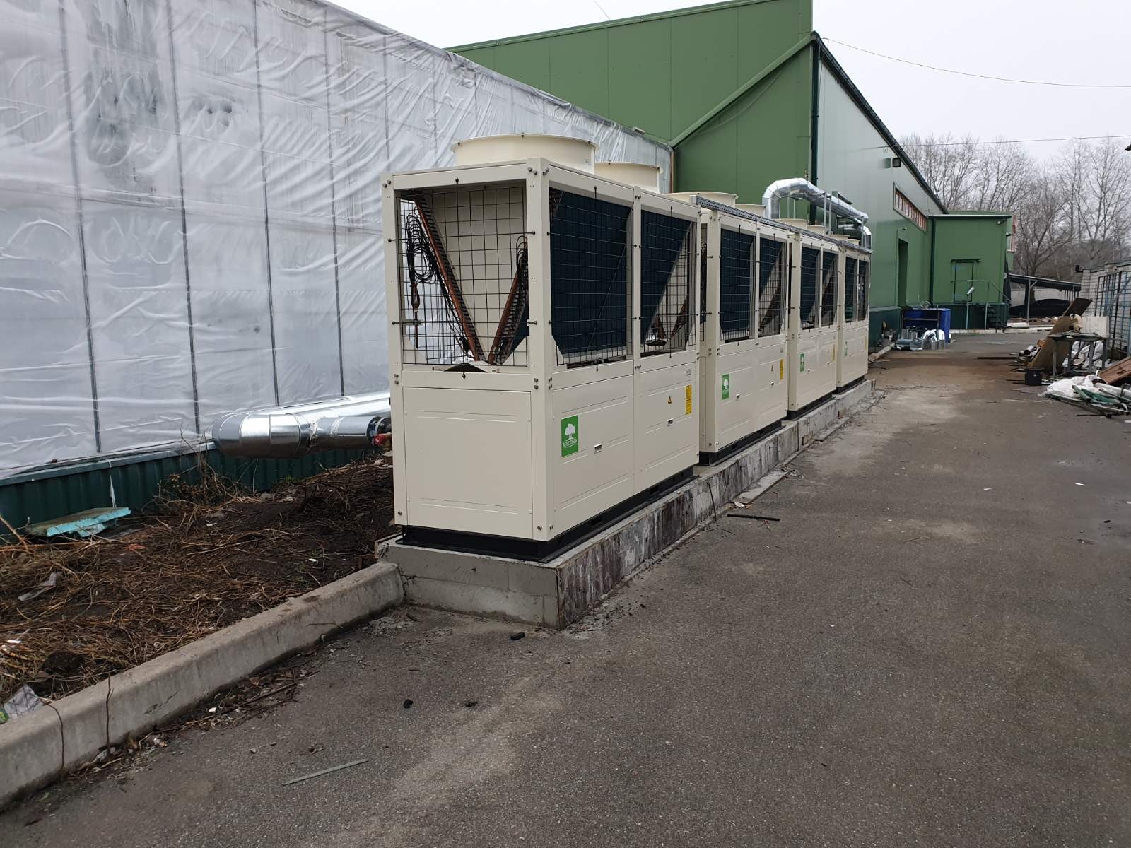 Szereg zewnętrznych jednostek wentylacyjnych z kratami i zielonym logo drzewa, zainstalowanych na betonowym fundamencie, z widoczną instalacją rur i budynkiem przemysłowym w tle.