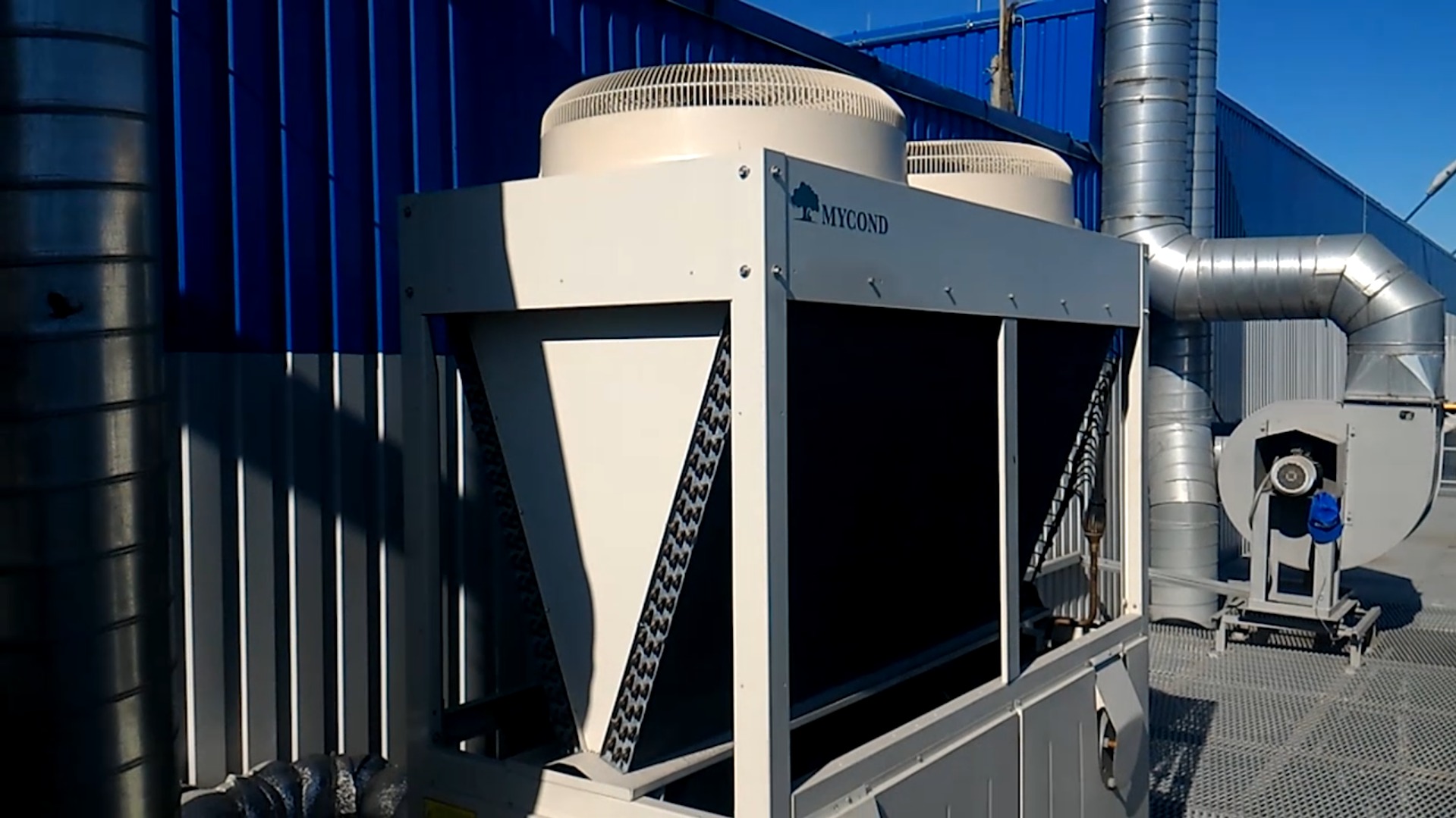 Biała jednostka zewnętrzna klimatyzacji przemysłowej marki Mycond z widocznymi wentylatorami i rurami wentylacyjnymi na tle niebieskiej ściany budynku.