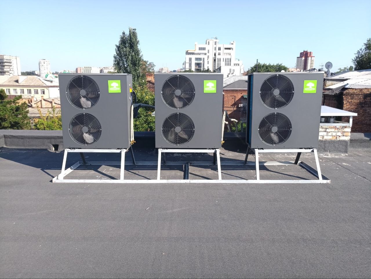 Trzy szare, dwuwentylatorowe jednostki zewnętrzne na dachu budynku, każda z zielonym logo w górnym rogu, ustawione na białych, metalowych stelażach na tle zabudowy miejskiej w słoneczny dzień.