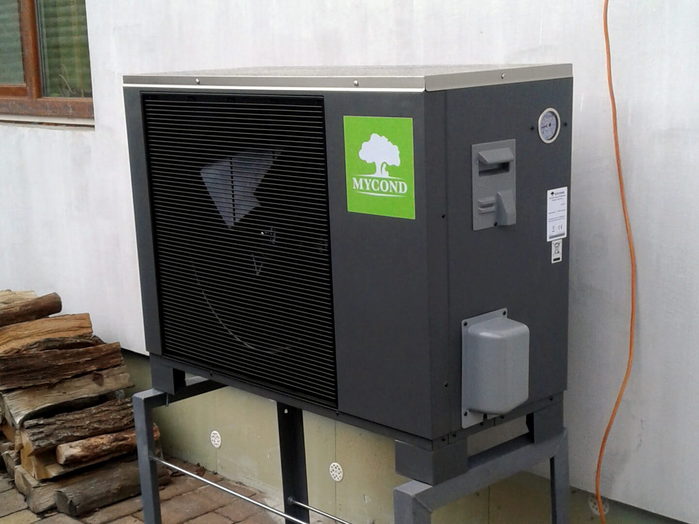 Szara pompa ciepła marki Mycond, zamontowana na metalowej konstrukcji, obok stos drewna na brukowanym podłożu, z pomarańczowym kablem biegnącym wzdłuż białej ściany budynku.