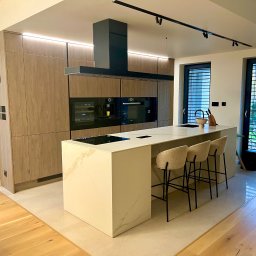 CUBE27 SPÓŁKA Z OGRANICZONĄ ODPOWIEDZIALNOŚCIĄ - Nowoczesna kuchnia z wyspą: fronty szafek z fakturą drewna, czarny okap, marmurowy blat wyspy z płytą indukcyjną, trzy beżowe hokery, widok na ogród przez okno z żaluzjami.