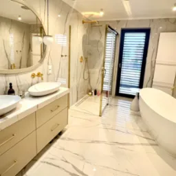 Elegancka łazienka z marmurowymi ścianami i podłogą, wyposażona w dwie białe umywalki nablatowe z złotymi bateriami, owalne lustro z oświetleniem, kabinę prysznicową ze złotymi detalami...
