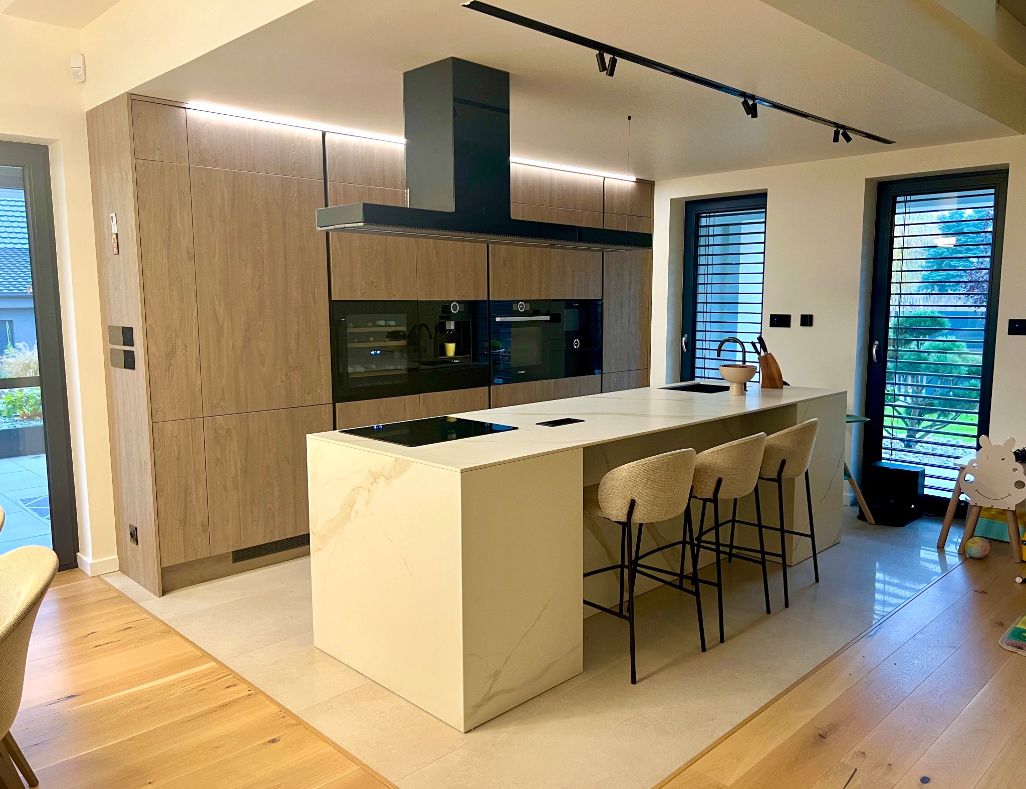 Nowoczesna kuchnia z wyspą: fronty szafek z fakturą drewna, czarny okap, marmurowy blat wyspy z płytą indukcyjną, trzy beżowe hokery, widok na ogród przez okno z żaluzjami.