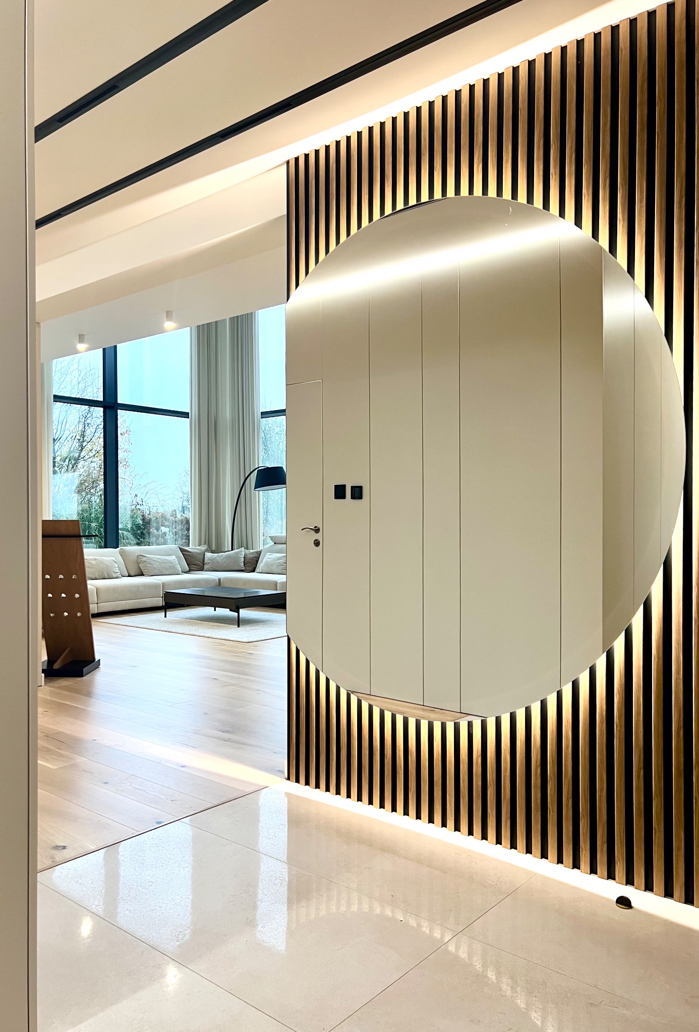 Nowoczesne wnętrze: wejście do salonu z okrągłym lustrem w obudowie z drewnianych listew z podświetleniem LED, widok na salon z dużym oknem i beżową sofą.