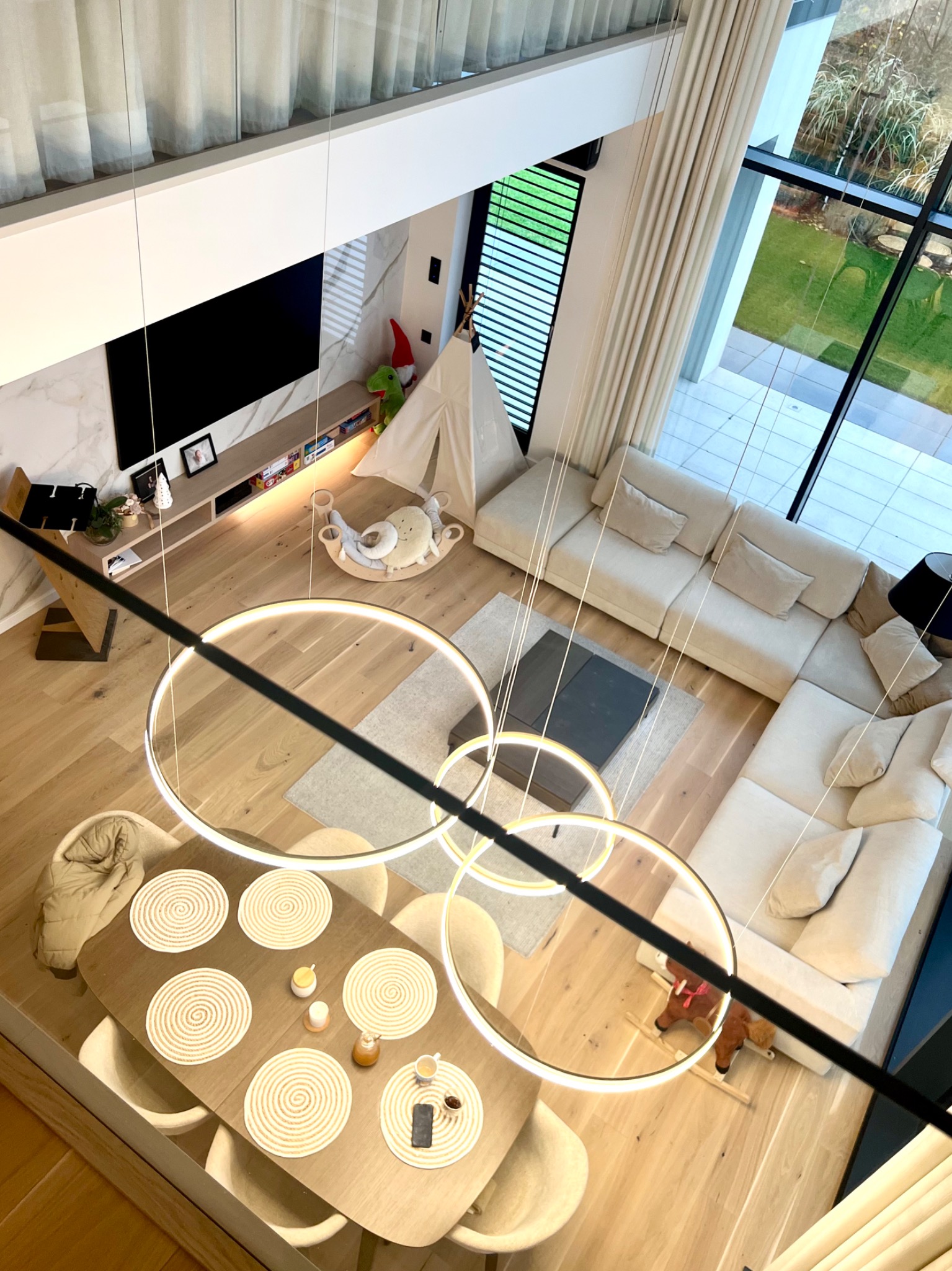 Wnętrze domu z widokiem z góry: jadalnia z nakrytym stołem, salon z narożną sofą, pokój dziecięcy z tipi i zabawkami, duże okna z widokiem na ogród, nowoczesne oświetlenie w postaci okręgów.