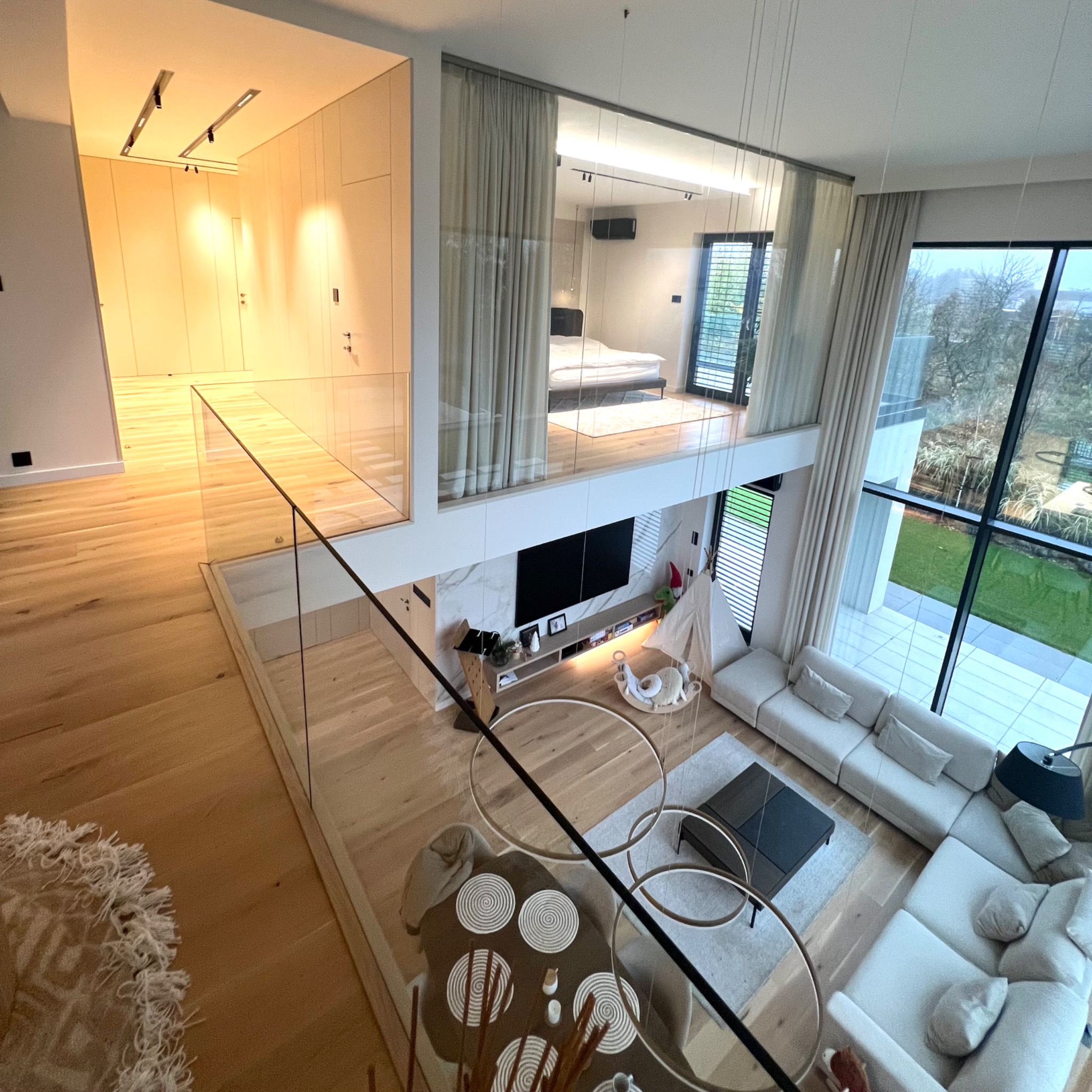 Dwupoziomowy salon z antresolą, widok z góry na minimalistyczny wystrój, jasne drewno na podłodze, biała sofa modułowa, okno panoramiczne i szklane balustrady, elementem dekoracyjnym jest wisząca...